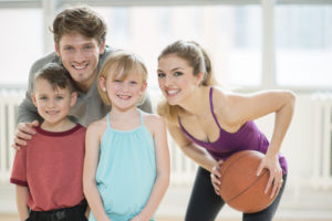A family of four are playing basketball together at the gym. They are smiling and looking at the camera.